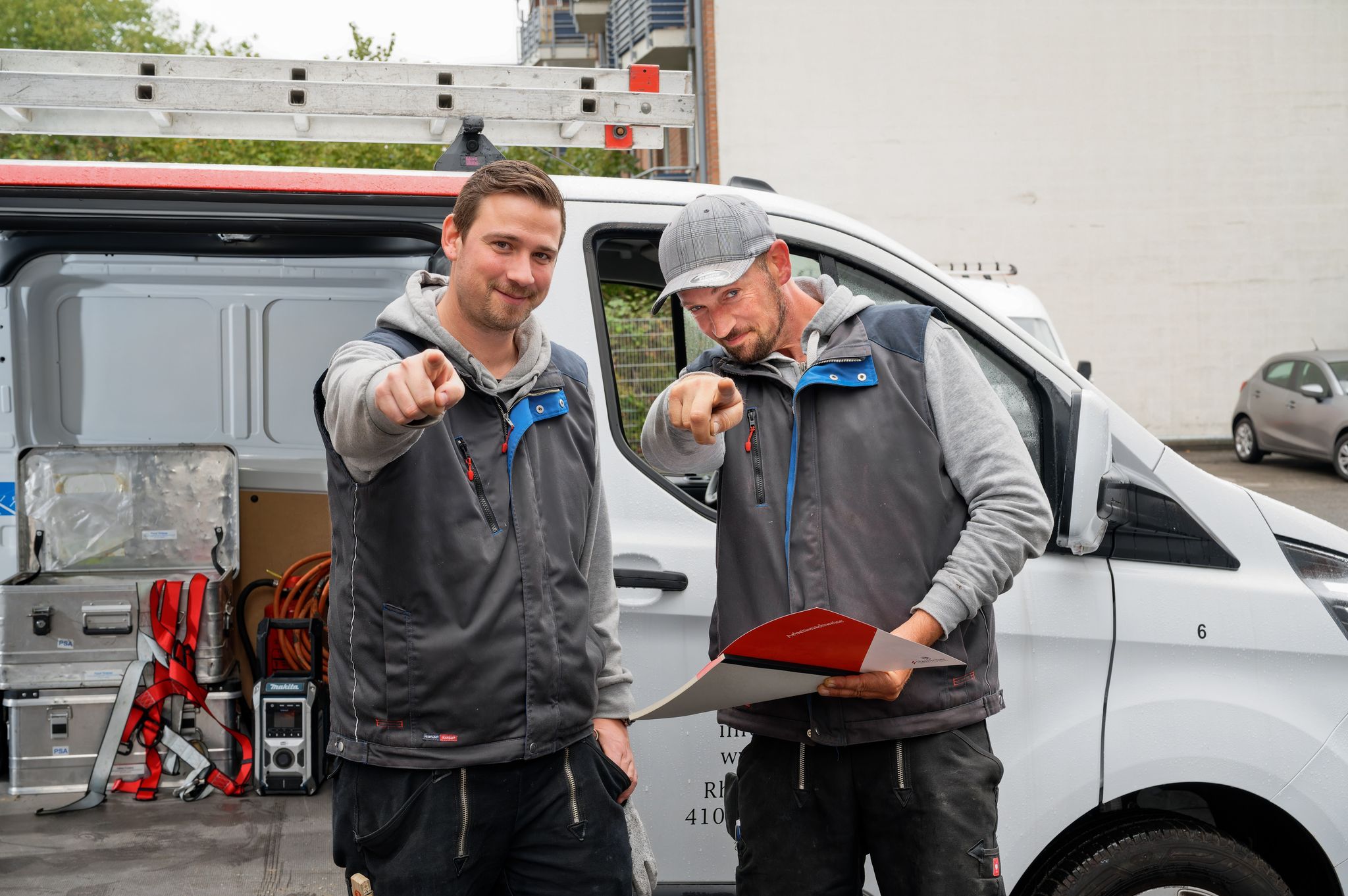 Zwei Mitarbeiter der Hamacher SolTec GmbH, die vor einem weißen Firmenfahrzeug stehen, mit den Zeigefingern in Richtung der Kamera zeigen und lächeln“.