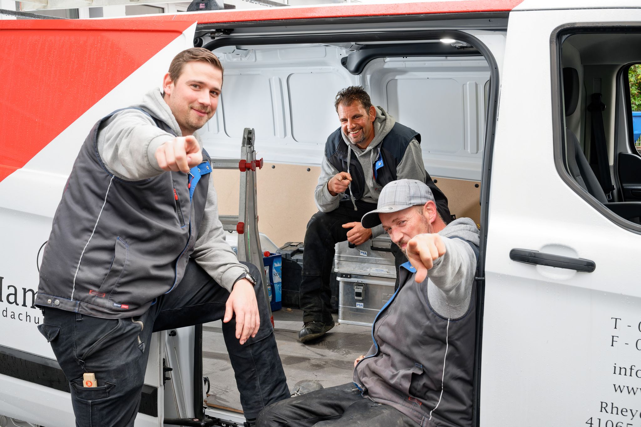 Drei junge Männer, Mitarbeiter der Hamacher SolTec GmbH, stehen am Firmenfahrzeug, lächeln in die Kamera und zeigen mit den Zeigefingern in die Kamera“.
